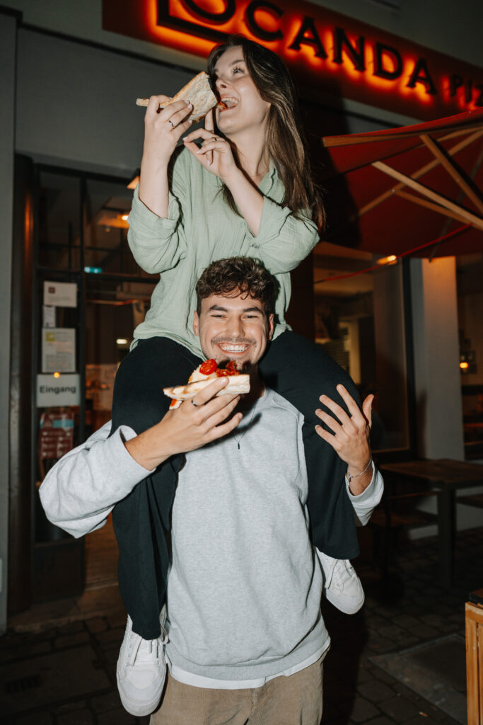 Junger Mann hat Freundin auf den Schultern, beide essen ein Stück Pizza vor der Locanda